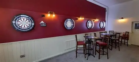 Four illuminated dart board setups on a red wall in a game room with seating.