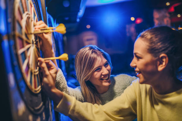 Smiling women enjoy playing darts, placing a dart into the dart board.