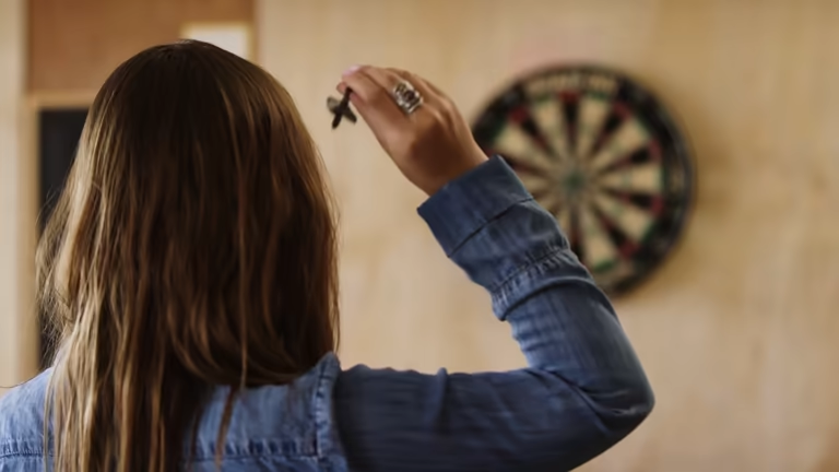 Woman from behind, aiming a dart towards the dart board on a wall.