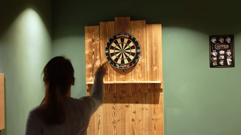 Woman throwing a dart at a dart board mounted on a wooden wall panel.
