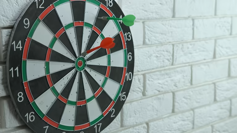 Two darts, red and green, stuck in a dart board on a white brick wall.