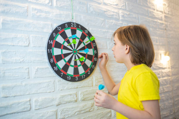 Child in yellow removing darts from a dart board on a white brick wall.